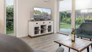 Ein Wohnzimmer mit Fernseher auf einer Kommode, einem Tisch mit Flasche und Gläsern sowie Blick auf einen Garten durch große Fenster.