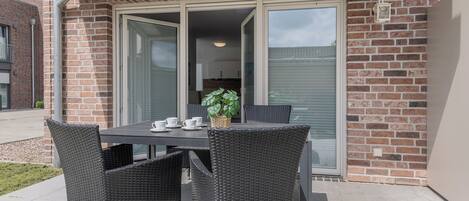 En udendørs terrasse med et bord, stole, en potteplante og en skydedør til et køkken.