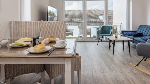 Ein modernes Wohnzimmer mit Essbereich, ausgestattet mit Rattanmöbeln, einem blauen Sofa und einem Balkon mit Blick auf Gebäude.