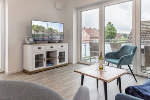 A modern living room with a television, a wooden table with a bottle and glass, and a balcony with wicker furniture. - Apartment (Carolinensiel)