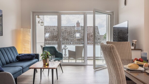 A modern living room with a sofa, armchair, and dining table. A large window offers a view of a traditional building with a tiled roof. - Apartment (Carolinensiel)