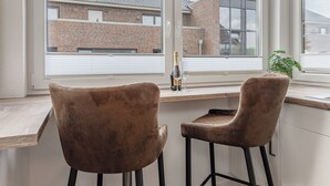 Et værelse med to brune stole, et træskrivebord og et stort vindue med udsigt til murstensbygninger.