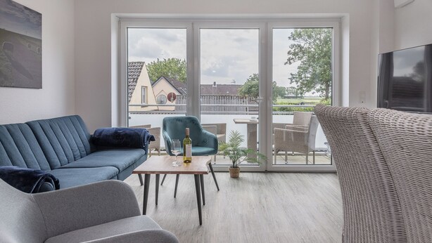 En moderne stue med en blå sofa, en grå lænestol og en fletstol. Der er et træbord med en flaske og et glas. Store vinduer giver udsigt udad.