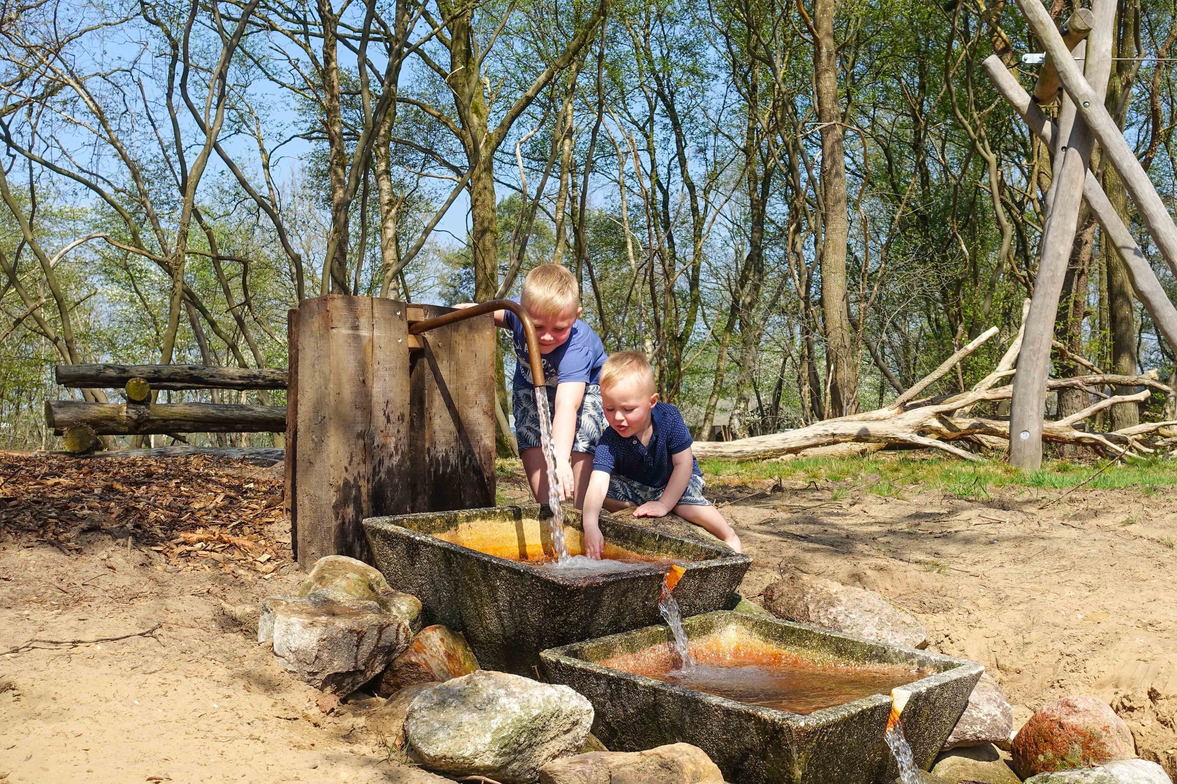 Two children playing with water from a rustic outdoor water feature surrounded by natural forest.