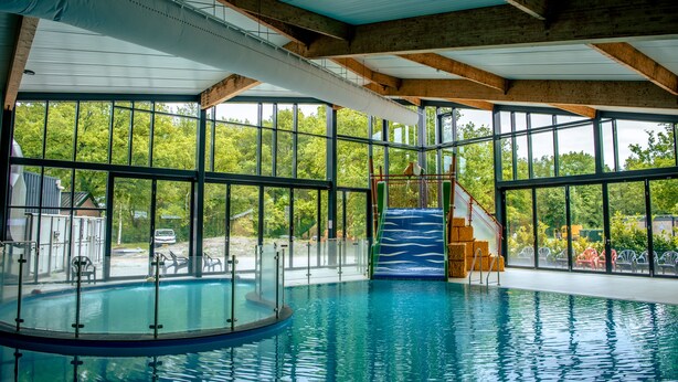 An indoor swimming pool with clear glass walls, a slide, and a view of the surrounding greenery. - EuroParcs Ruinen (Ruinen)