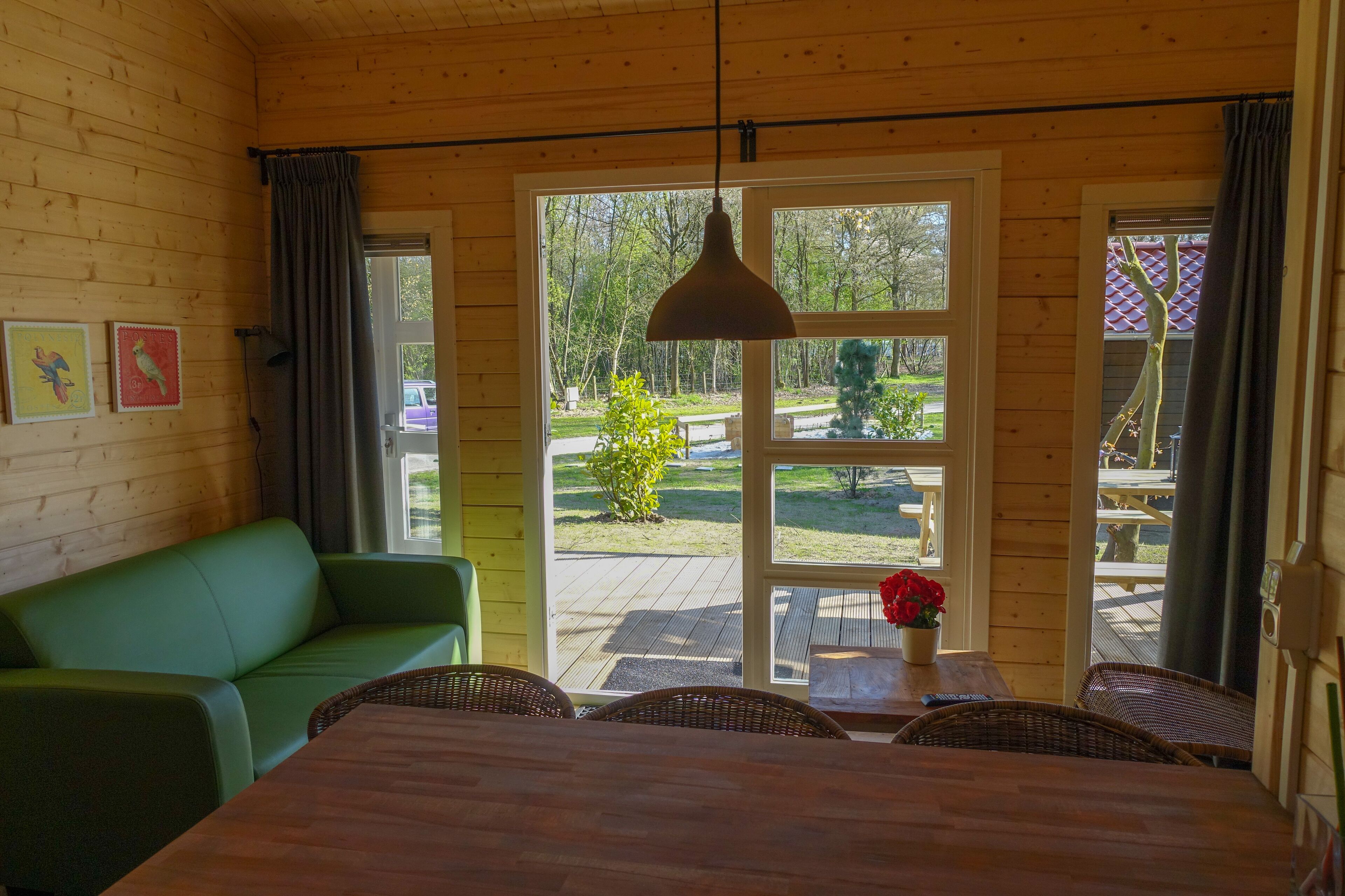 A wooden cabin interior with a dining area, green sofa, wicker chairs, and a view of a garden through large windows.