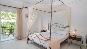A four-poster bed with a canopy, two bedside tables with lamps, a balcony with a table and chairs, and a view of greenery outside. - Haris Villas (Viannos)