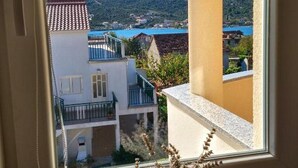 Una ventana con vista a un pueblo costero, un balcón y un pequeño jarrón con flores secas.