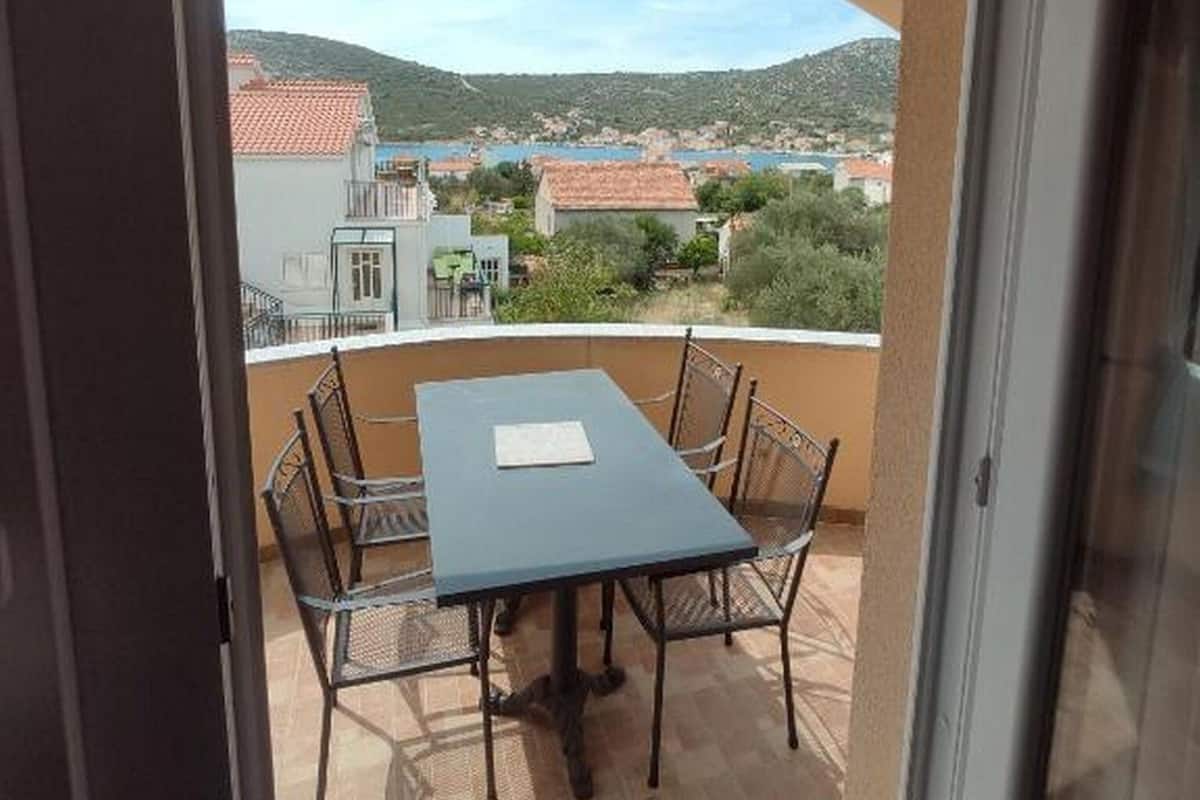 Un balcon avec une table et des chaises, offrant une vue sur une ville avec des bâtiments aux toits rouges et un plan d’eau.