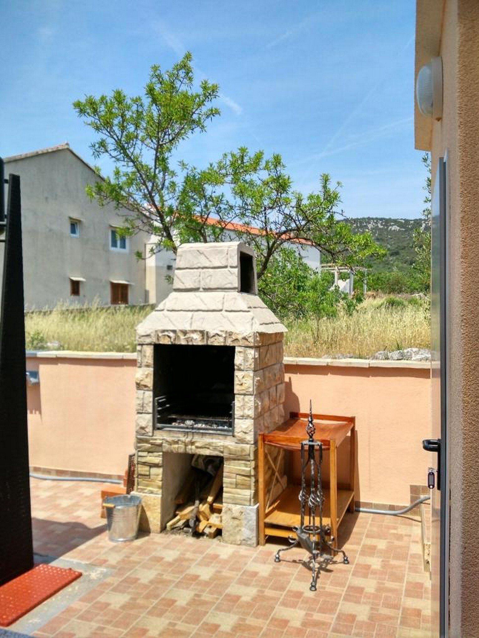 Un'area barbecue esterna in pietra con un camino, un tavolo in legno e un supporto per griglia in metallo.