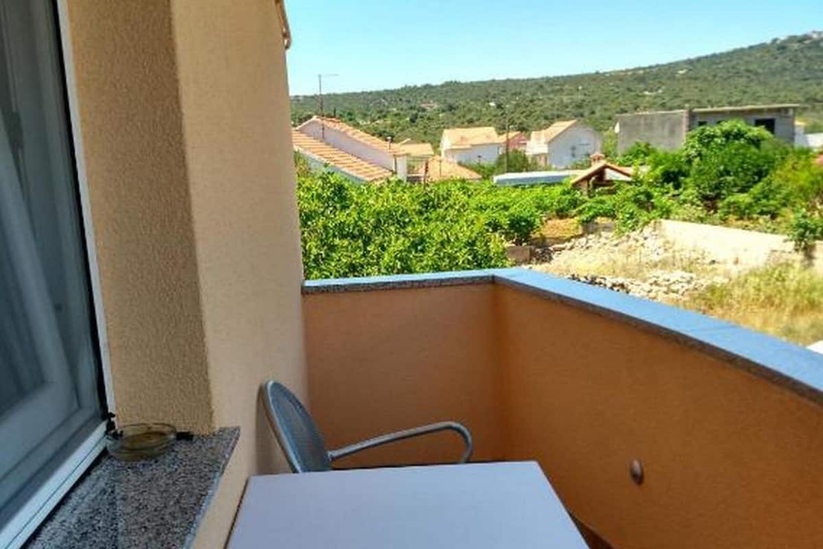 Un balcon avec une table et deux chaises, une fenêtre donnant sur des immeubles et de la verdure, et une porte avec un paillasson.