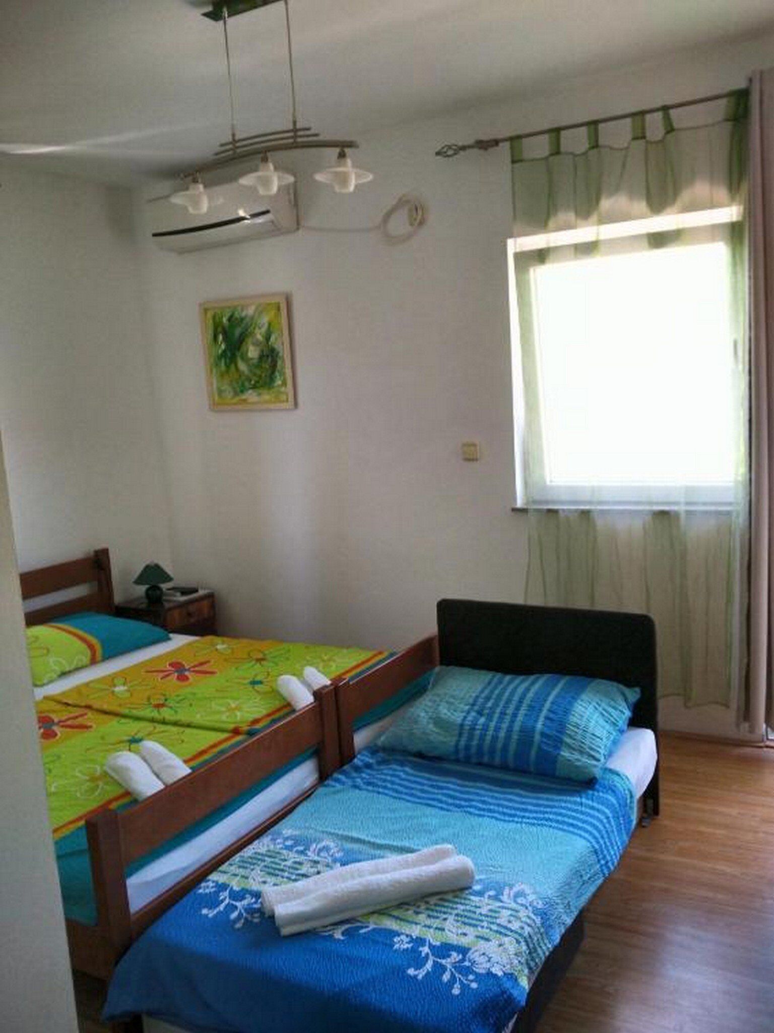 A room with two single beds, a ceiling fan, a window with curtains, and a painting on the wall.