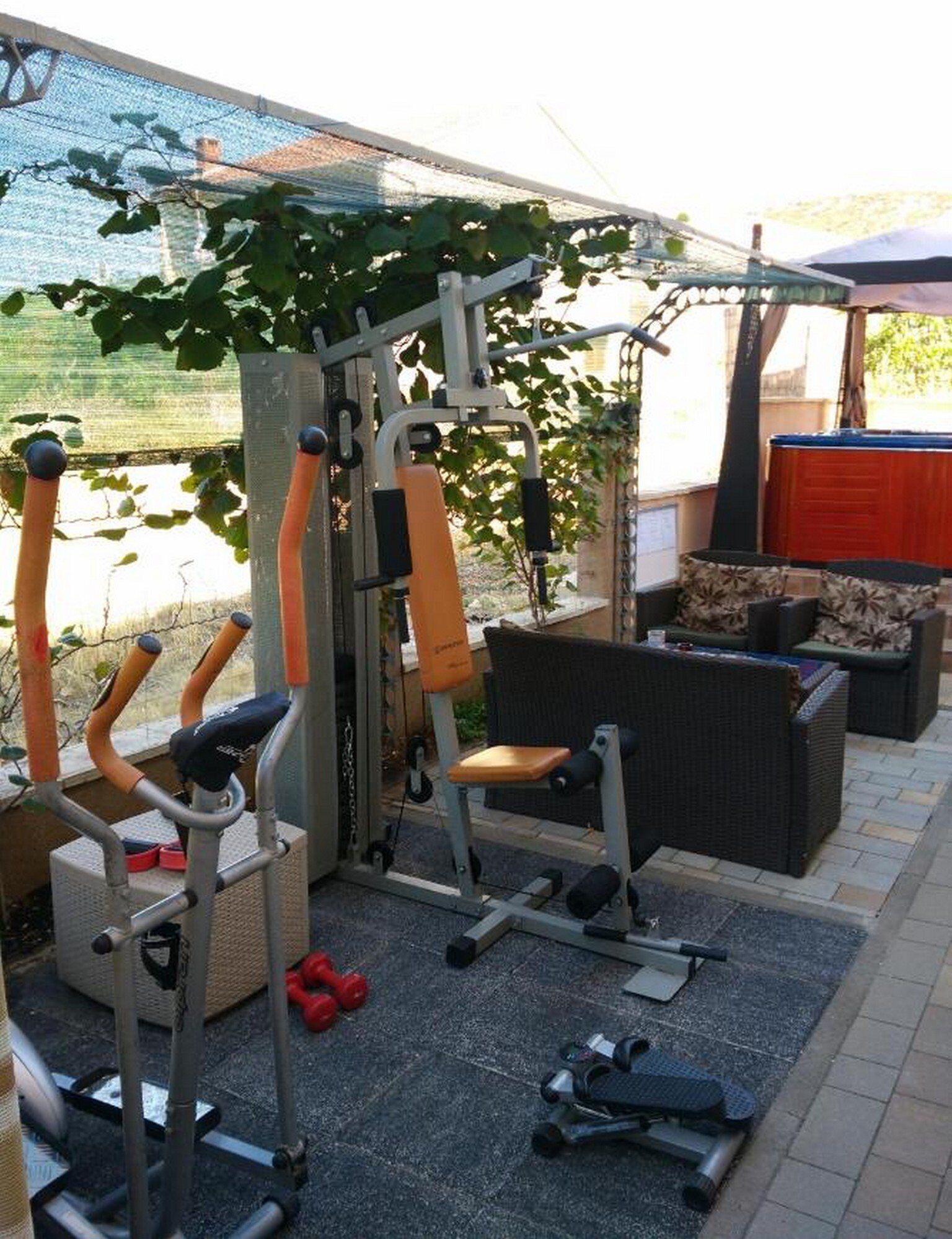 Ein Outdoor-Fitnessbereich mit Trainingsgeräten, einer überdachten Sitzgelegenheit und Blick auf einen Garten.