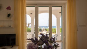 A room with a television, a vase of flowers, and a view of an outdoor patio with a table and chairs. - VILLA SUNSET - Seafront (Torre Suda)