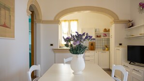 A dining area with a white table, white chairs, a white vase with flowers, and a kitchen in the background. - VILLA SUNSET - Seafront (Torre Suda)