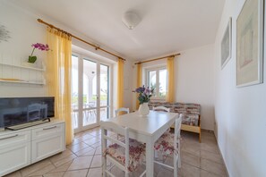 Quarto com televisão sobre um armário branco, mesa de jantar com cadeiras, um sofá e janela com cortinas amarelas.