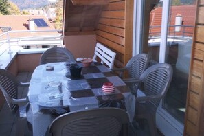 A balcony with a checkered tablecloth, white chairs, and a small table with a bowl and a red and white striped cup.