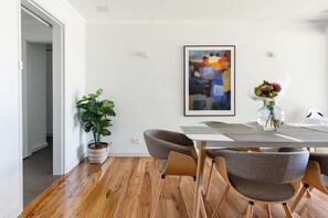A dining area with a wooden floor, a white table, and four grey chairs. There is a potted plant and a framed abstract painting on the wall.