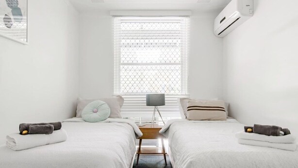 Ein schlicht eingerichtetes Schlafzimmer mit zwei Betten, einem Fenster mit Jalousien, einer Nachttischlampe und einer Klimaanlage.
