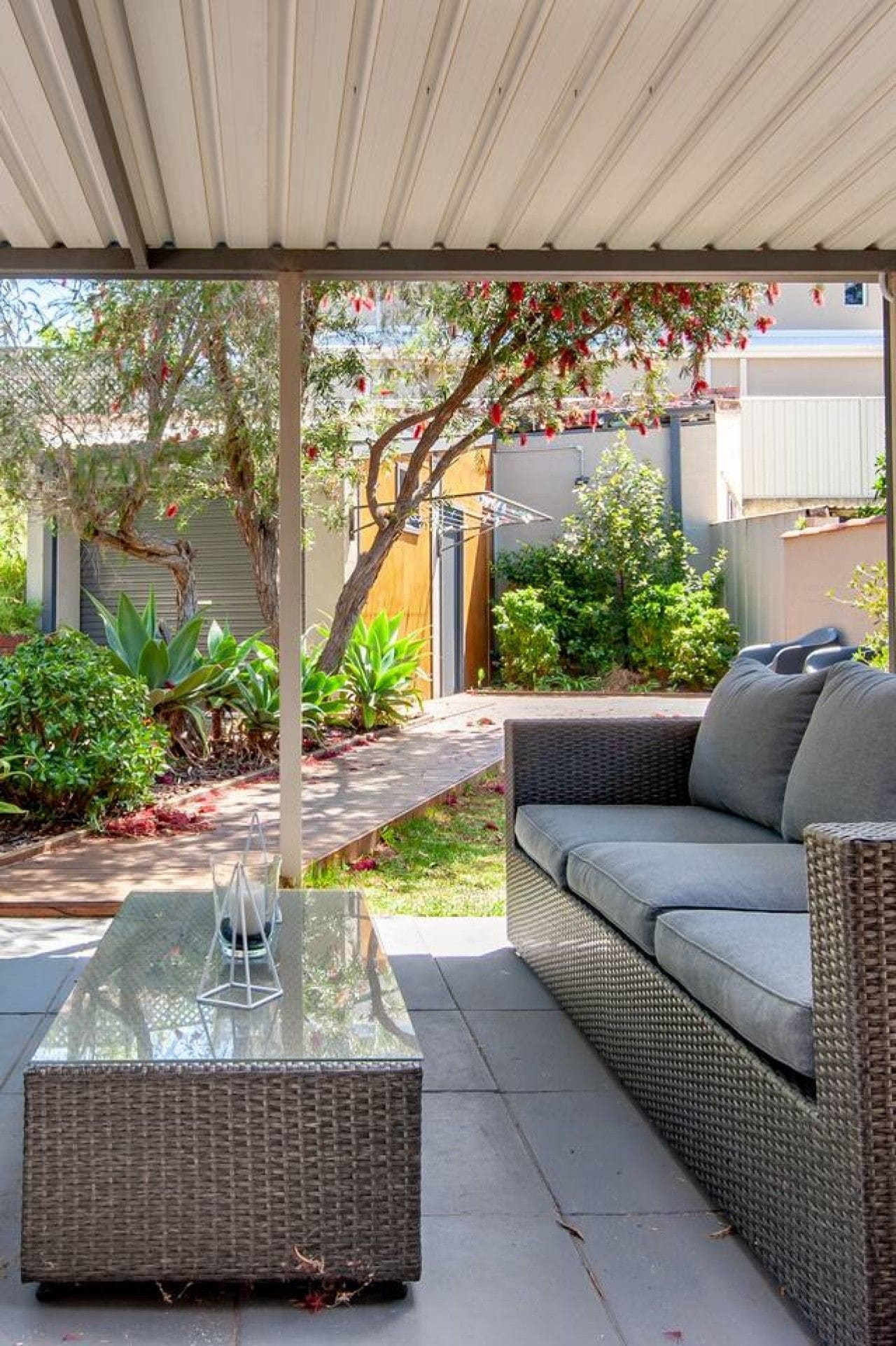 Une terrasse extérieure dotée d’une table à plateau de verre, de meubles en osier et d’un espace abrité offrant une vue sur un quartier résidentiel.