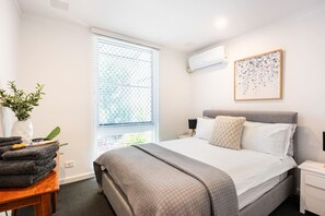 A hotel room with a bed, bedside table, air conditioning unit, window with blinds, and a framed artwork on the wall.