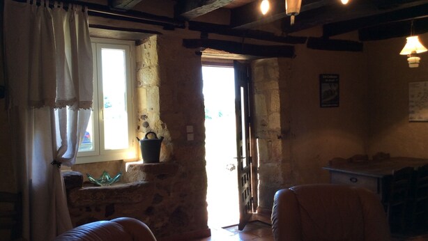 Una habitación rústica con pared de piedra, techo de madera y ventana con cortinas. Hay una puerta que da al exterior, una mesita y dos sillas.