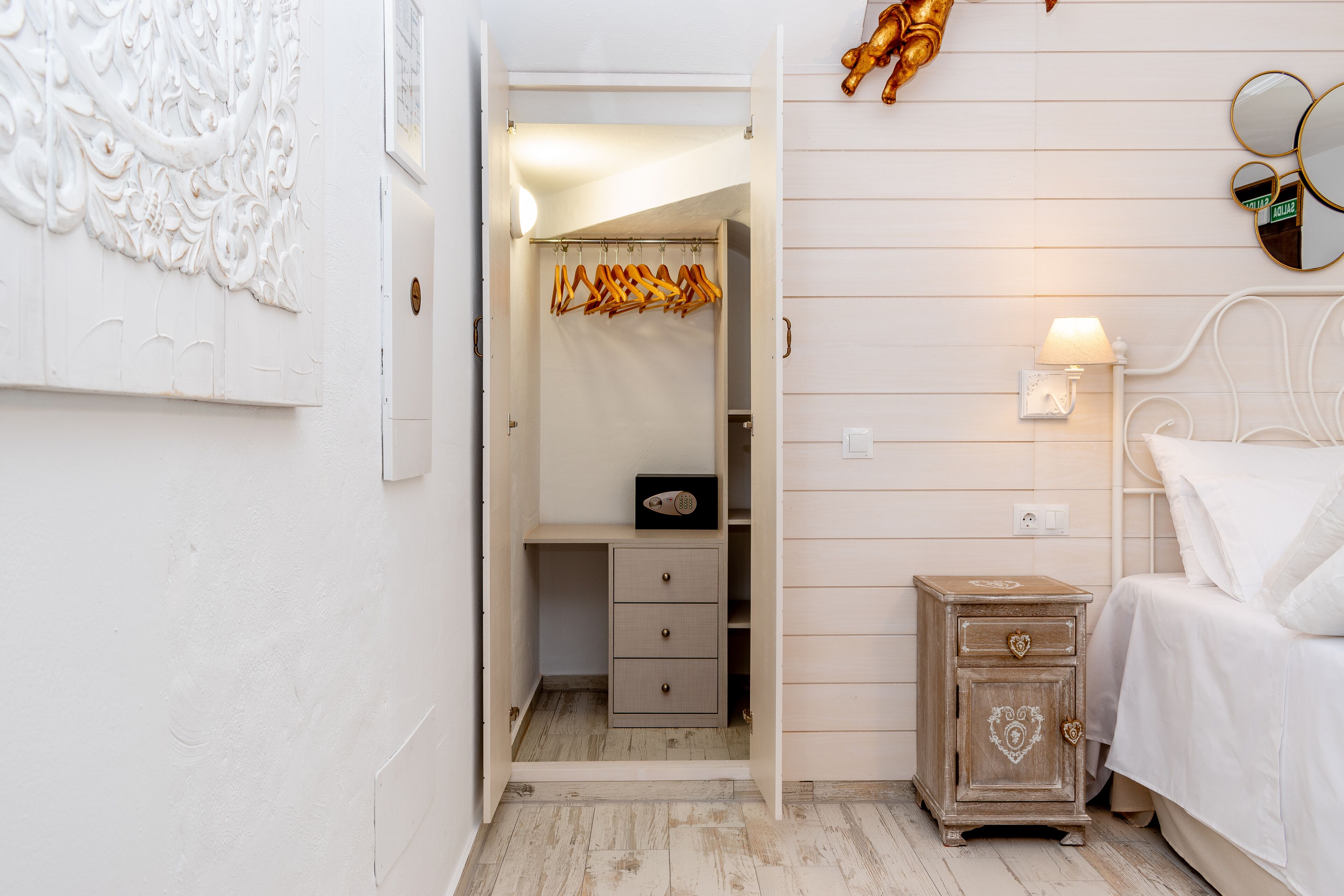 Une chambre avec une table de chevet en bois, un lit avec une literie blanche, une lampe de chevet et une armoire où sont suspendus des vêtements.