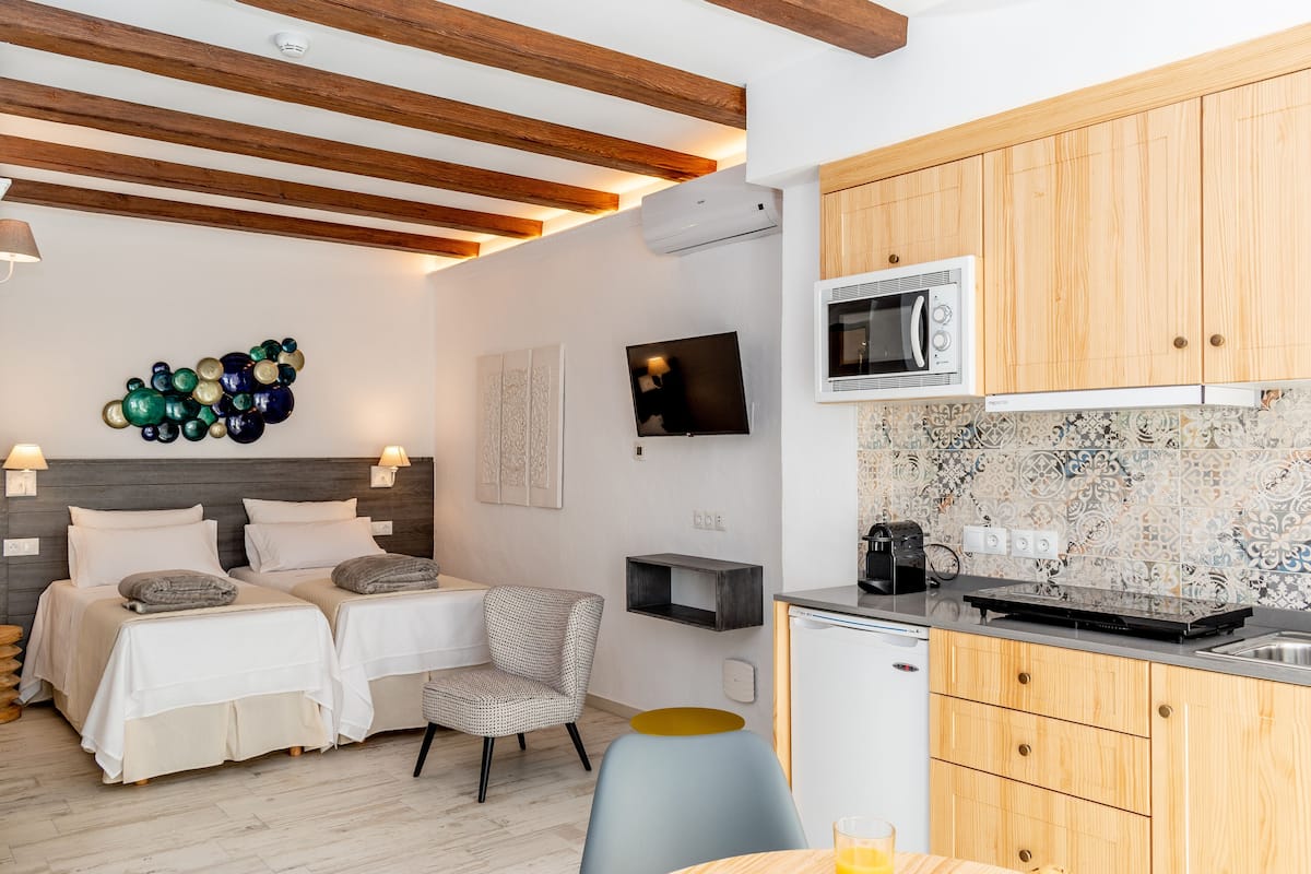 A modern bedroom with two beds, a TV, and a kitchenette with wooden cabinets and a microwave.
