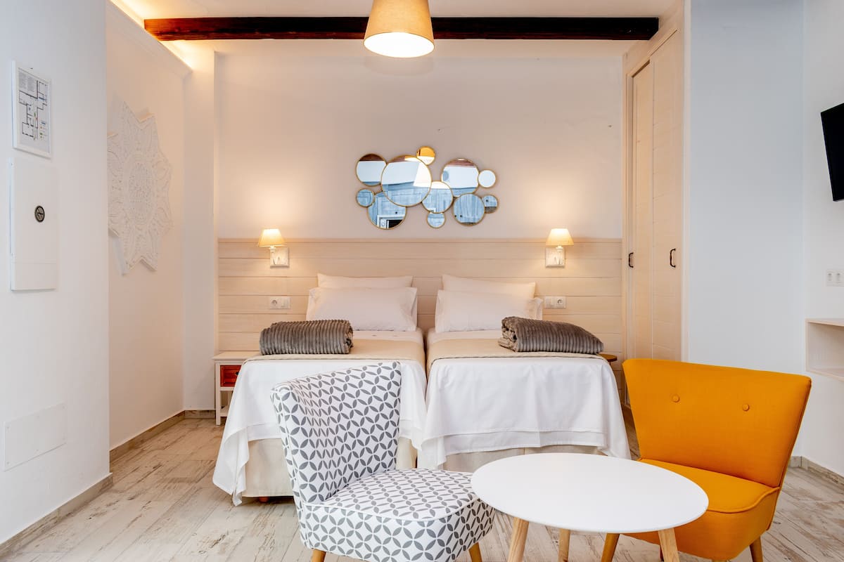 A modern hotel room with two beds, a round table, two patterned chairs, and a unique wall art piece.