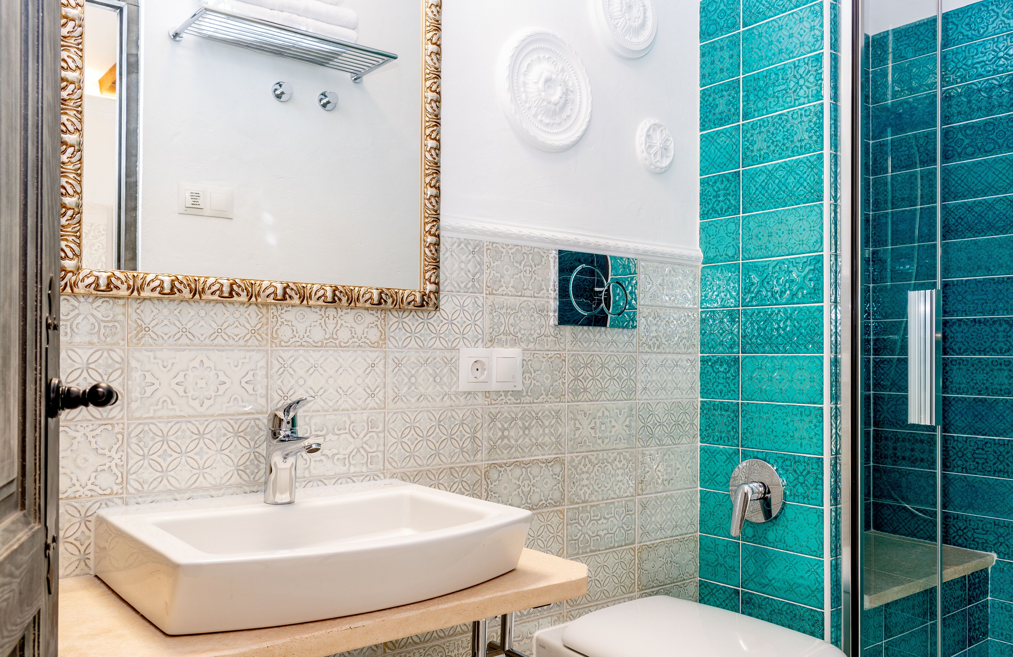 Een badkamer met een wit wastafelmeubel, een spiegel met een sierlijk lijstwerk en een douchegedeelte met blauwe tegels.