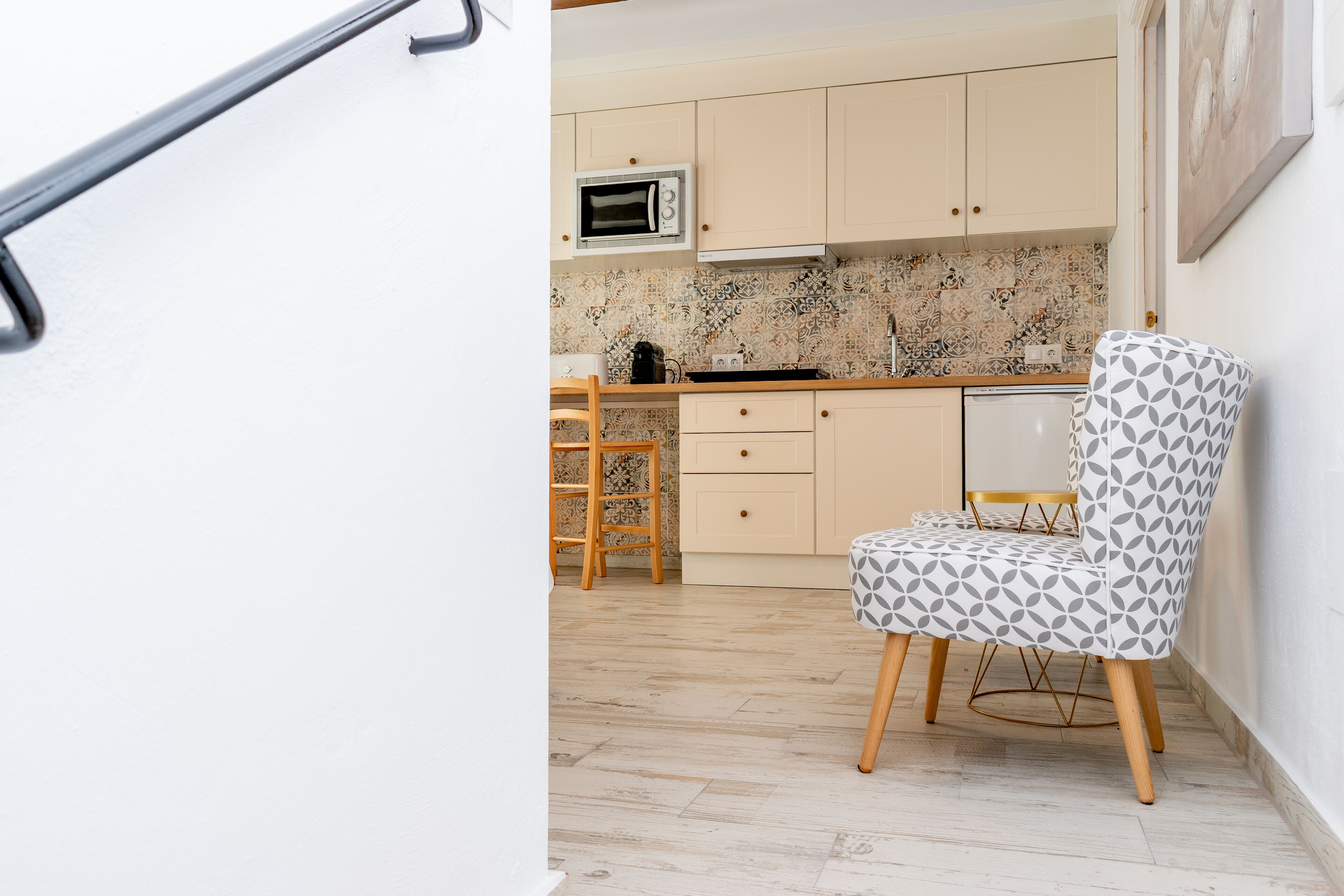 Una cocina con armarios color beige, un microondas, una silla de madera y un otomano estampado.