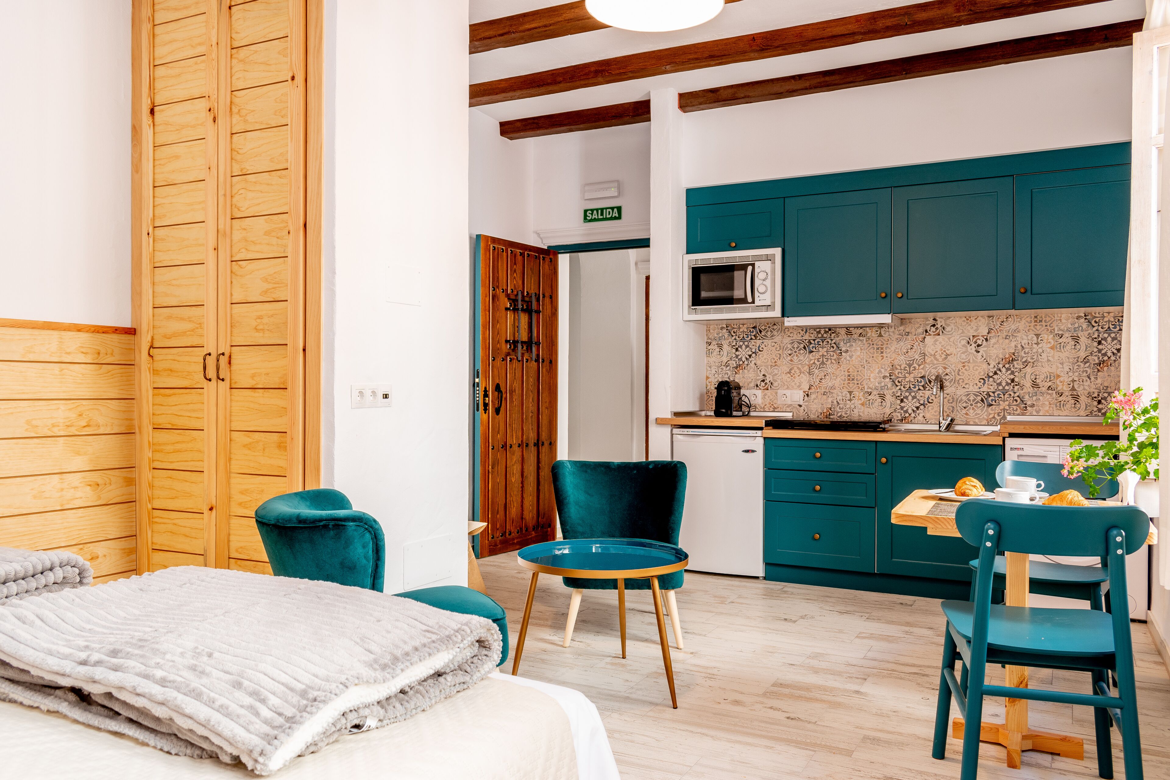 A room with a bed, a teal chair, a small table, and a kitchenette with teal cabinets and a microwave.