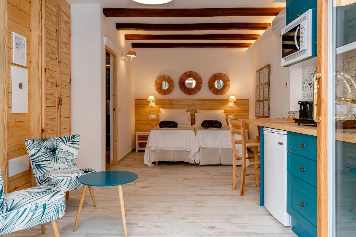 A compact room with a bed, two bedside tables, a small table, and a kitchenette.