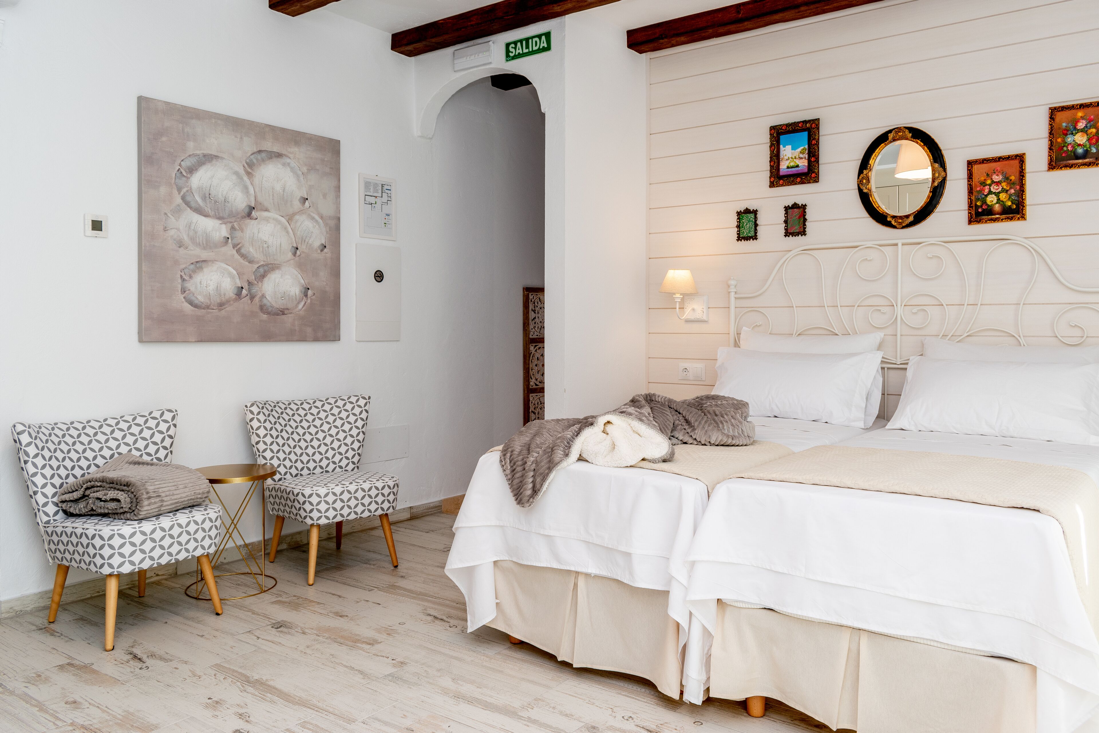 A bedroom with a bed, two patterned chairs, a small table, and artwork on the wall.
