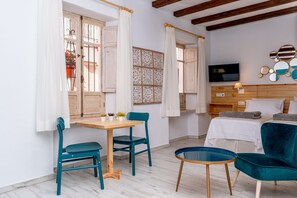 A room with a wooden table, two teal chairs, a small round table, a bed with a white bedspread, a TV mounted on the wall, and a decorative mirror arrangement.