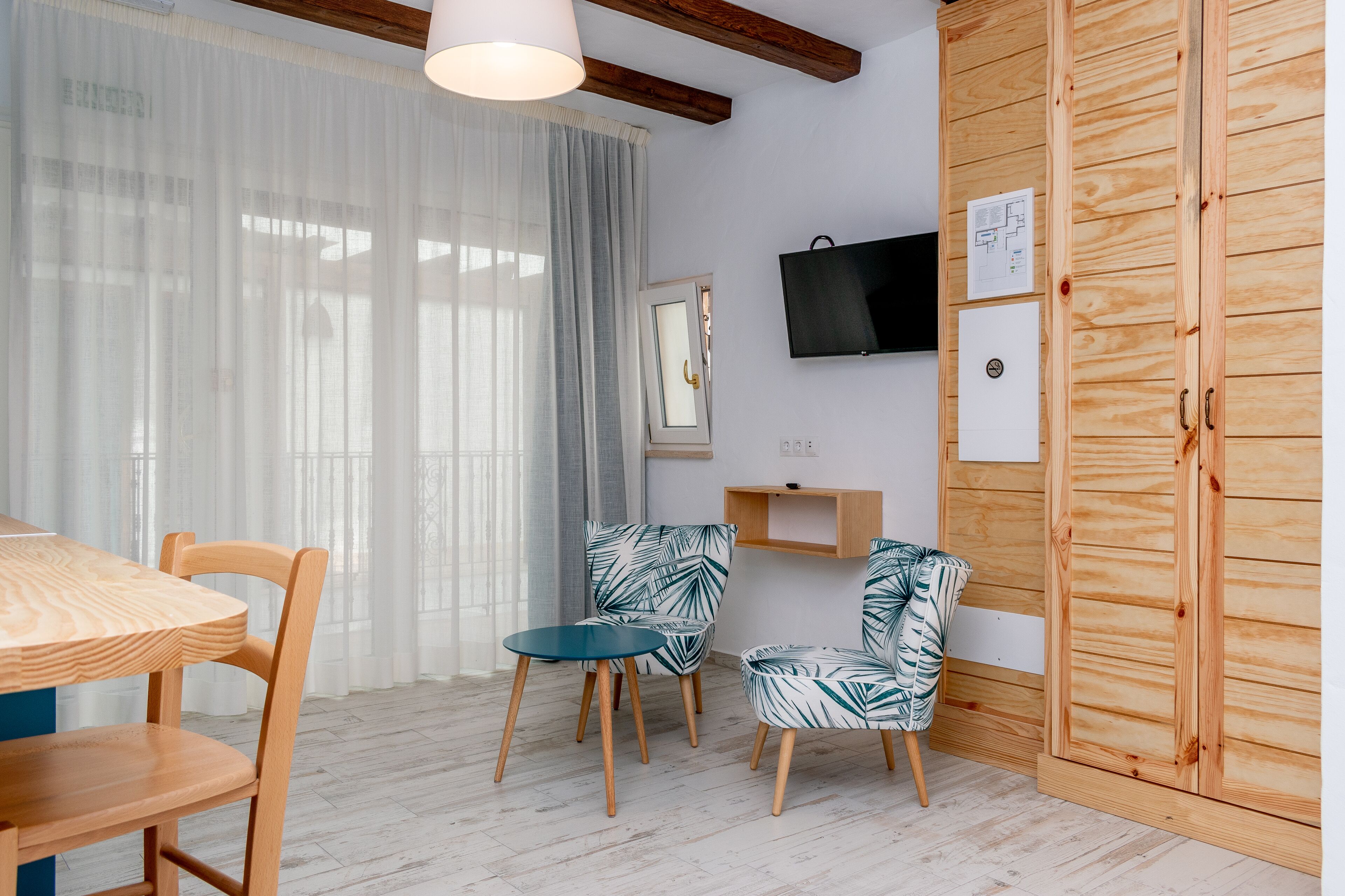 A room with a wooden dining table, two chairs, a wooden wardrobe, a television, and a small shelf.