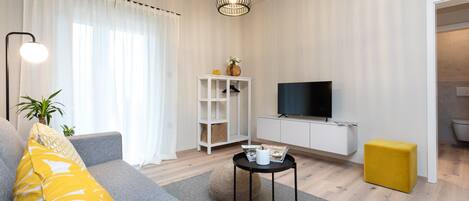 A modern living room with a grey sofa, a yellow ottoman, a black coffee table, and a flat-screen TV on a white wall.