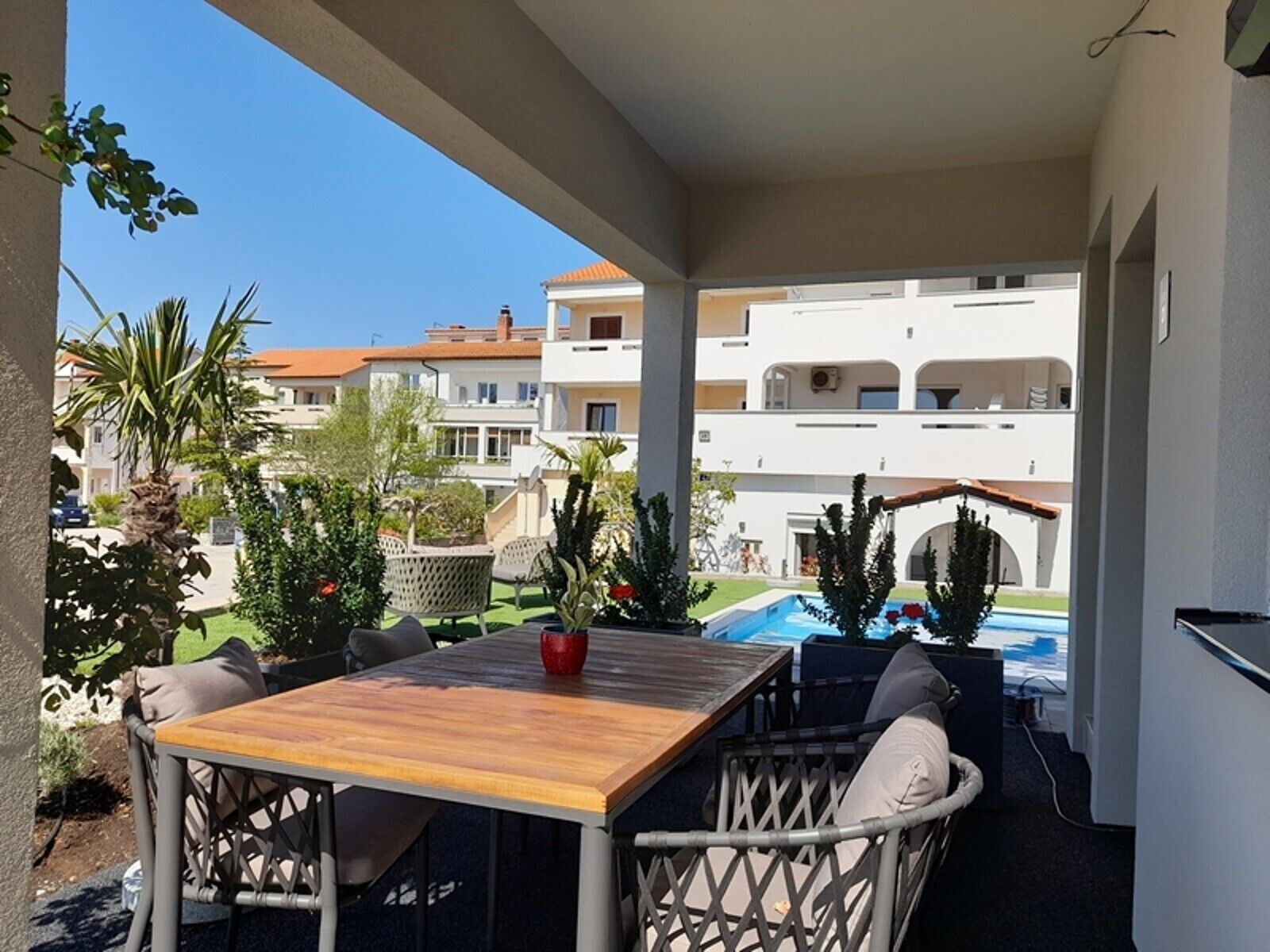An outdoor patio with a wooden table and wicker chairs, overlooking a swimming pool and neighboring buildings.