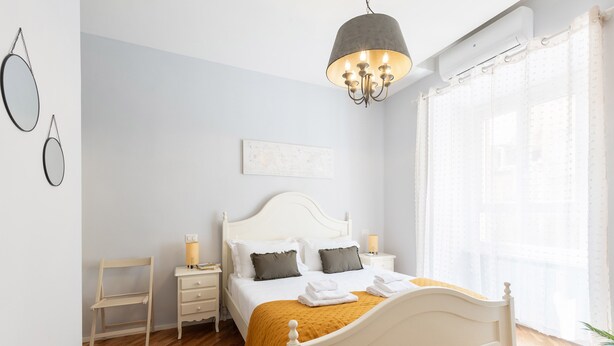 A bedroom with a white bed, a wooden chair, a bedside table, and a hanging light fixture.
