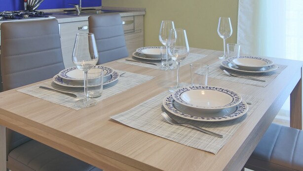 Una mesa de comedor dispuesta con platos, vasos y cubiertos, con una cocina al fondo.