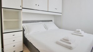 Ein ordentlich bezogenes Bett mit weißen Leinen und Handtüchern, ein grauer Kopfteil und ein weißer Kleiderschrank mit mehreren Schubladen.