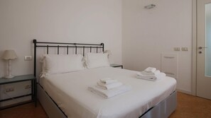 Cama bem arrumada com roupa de cama e toalhas brancas, mesa de cabeceira com abajur e luminária na parede.