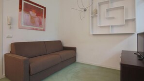 Uma sala de estar com um sofá marrom, uma TV fixada na parede, uma prateleira com objetos decorativos e um quadro.