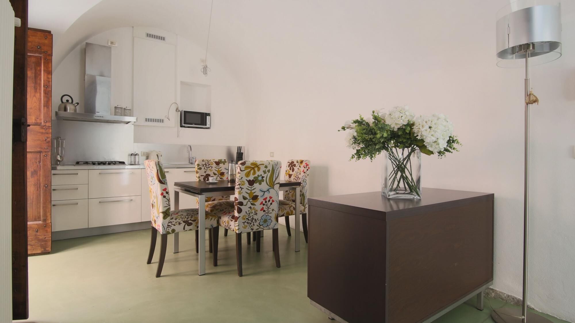 A compact kitchen with white cabinets, a microwave, and a gas stove. There is a dining area with floral-patterned chairs and a small table. A vase with white flowers is on a brown side table.