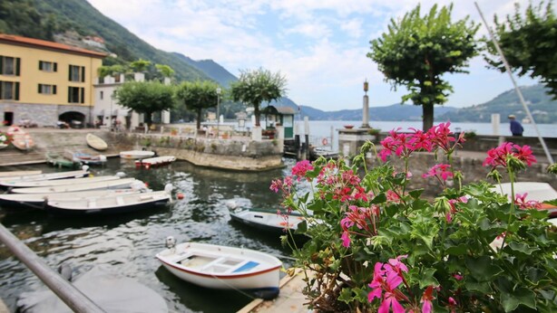 A lakeside dock with several small boats, a yellow building, and a mountain in the background. - Bartolomeo de Benz - Torno (Torno)