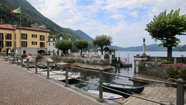 A waterfront area with boats docked, a stone staircase leading to a building, and a flagpole with the Italian flag.