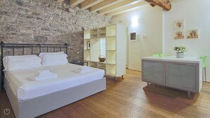 A bedroom with a stone wall, a bed with white linens, a wooden floor, a desk with a plant, and a green chair.