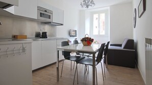 Ett modernt kök med vita skåp, en inbyggd ugn och en kaffemaskin. Det finns ett matbord med stolar och en korg med blommor.