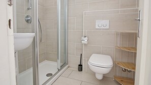 Ein Badezimmer mit einer gläsernen Duschkabine, einem weißen Waschbecken und einer Toilette. An der Wand befinden sich zwei Regale mit Holztüren und ein Toilettenpapierhalter.