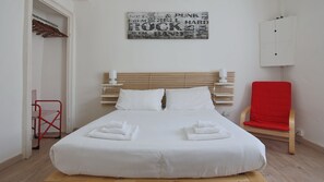 Una cama bien tendida con sábanas y toallas blancas, un cabecero de madera, una silla roja y un aplique de pared.